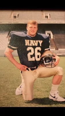 Tim Cannada kneels on the field in his number 26 Navy uniform, holding his helmet.